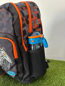 A black and gray patterned backpack with bright orange zippers and accents, shown on artificial grass with a blue water bottle in its side mesh pocket. The brand name 'Spikal' is visible on a blue tag near the bottom left.