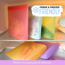 Five reusable silicone food sandwich and snack bags in red, blue, green, yellow and purple shown inside a refrigerator shelf, with text overlays reading 'FRIDGE & FREEZER FRIENDLY' and 'Perfect for meal prep & leftovers.'.