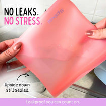 A red silicone leakproof sandwich and snack bag filled with water being held by two hands in a kitchen sink setting, with text overlays reading 'NO LEAKS. NO STRESS.