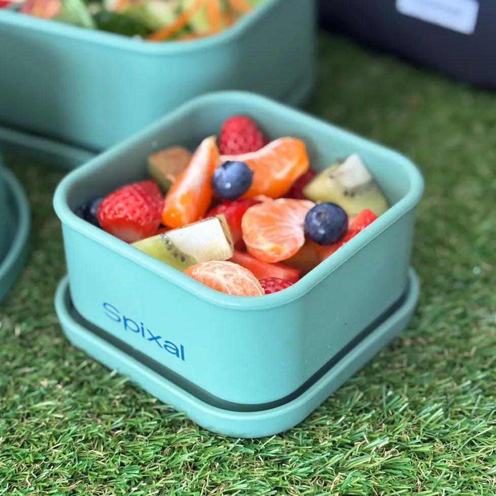 A green square lunch box with the brand name 'spixal' printed on the side, filled with fresh fruit including strawberries, blueberries, oranges, and kiwi, set on artificial green grass.